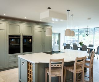 A spacious kitchen with a large sage green island featuring a built-in hob. The island has a curved white worktop, wooden bar stools, pendant lights, and a discreet ceiling extractor.