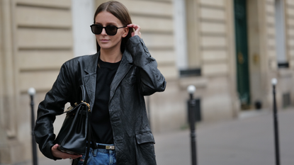 Diane Batoukina wears sunglasses, a black top, a black leather oversized blazer jacket, a Hermes belt, blue denim wide-leg flared jeans / pants, a Hermes bag, during a street style fashion photo session, on March 17, 2023 in Paris, France. 