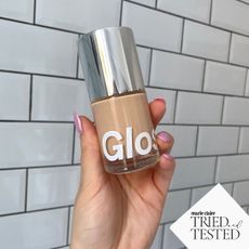 A hand holding Glossier Stretch Fluid Foundation up against some white bathroom tiles