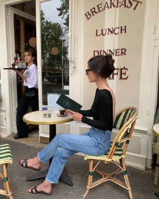 French influencer @leasy_inparis wearing a black long sleeve top, blue straight-leg jeans, and black open-toe sandal mules at a café in Paris