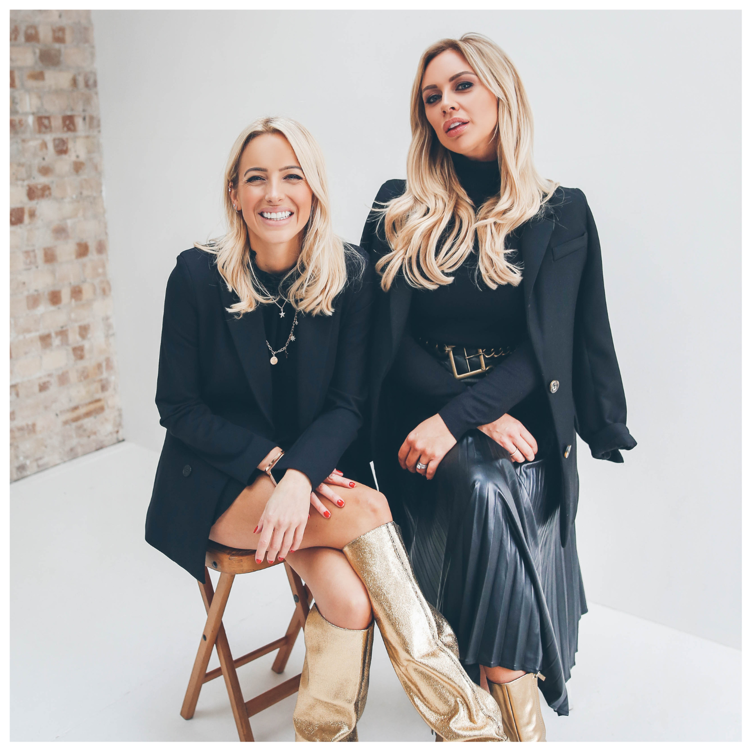 Two blonde girls in black coats and skirts with gold boots sitting beside each other on wood stools