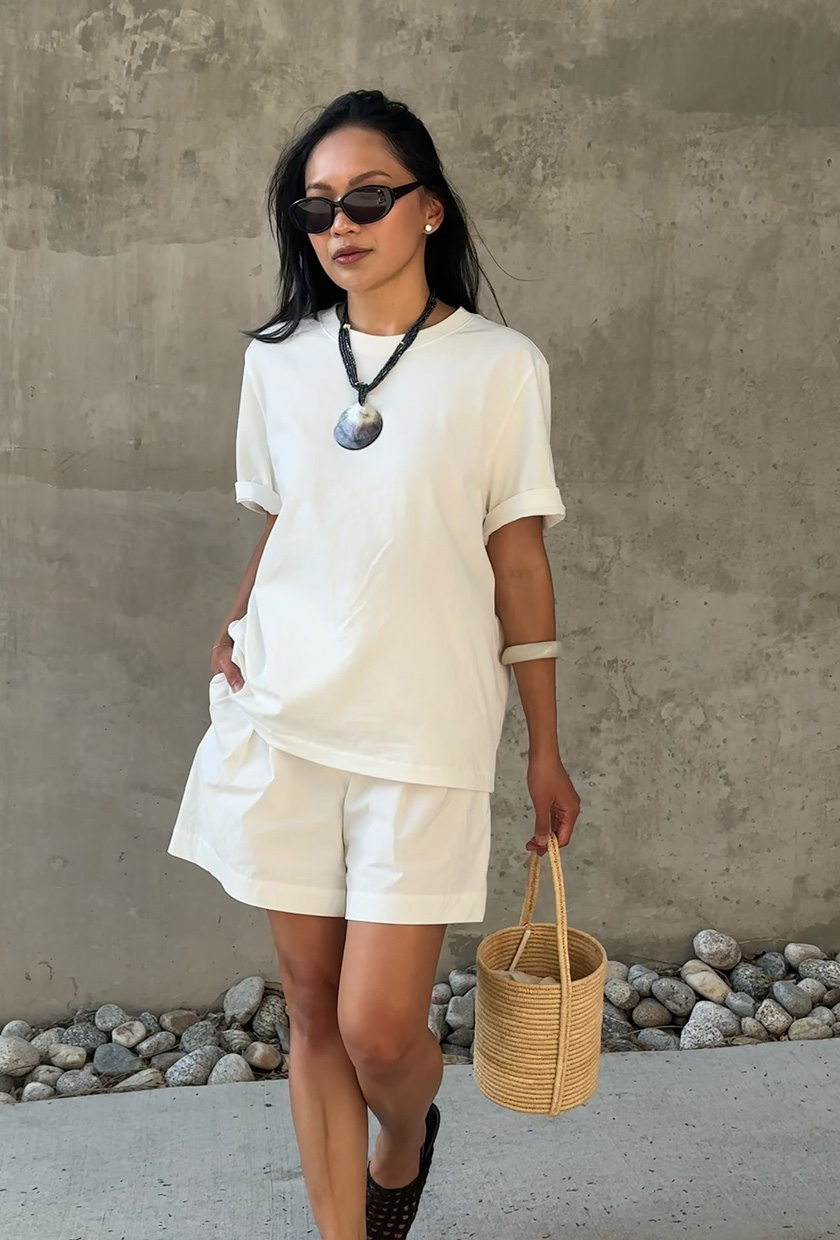 The coastal boho trend is shown in a street style photo of a woman standing on the sidewalk wearing a white t-shirt with white linen shorts styled with black sunglasses, a large black shell necklace, a white resin bangle, a raffia bucket bag, and black ballet flats