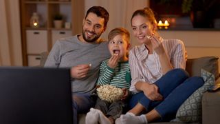 Man, woman and child watching TV together. All are smiling and eating popcorn, which the child is holding