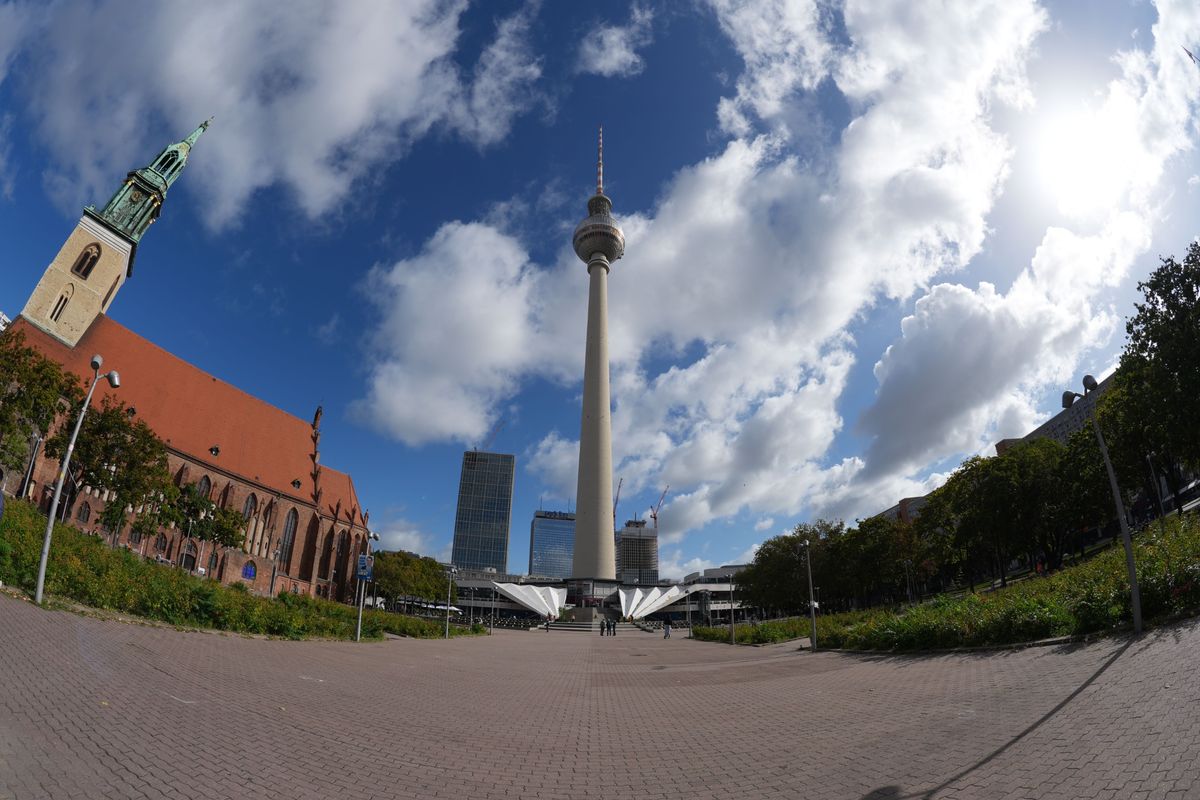 Ultra-wide view of Berlin&rsquo;s Fernsehturm TV tower and St. Mary&rsquo;s Church beneath a dramatic blue sky with large white clouds.