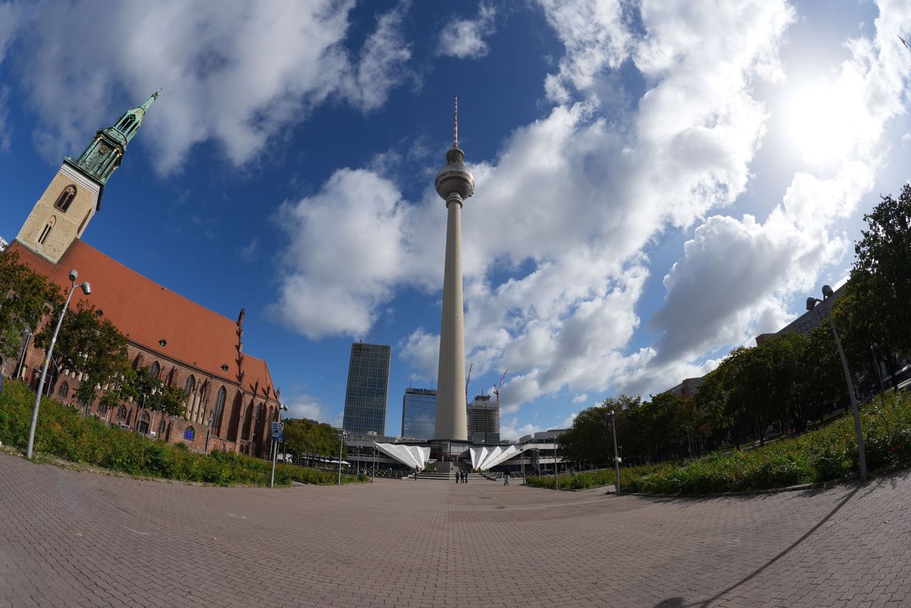 Ultra-wide view of Berlin&rsquo;s Fernsehturm TV tower and St. Mary&rsquo;s Church beneath a dramatic blue sky with large white clouds.