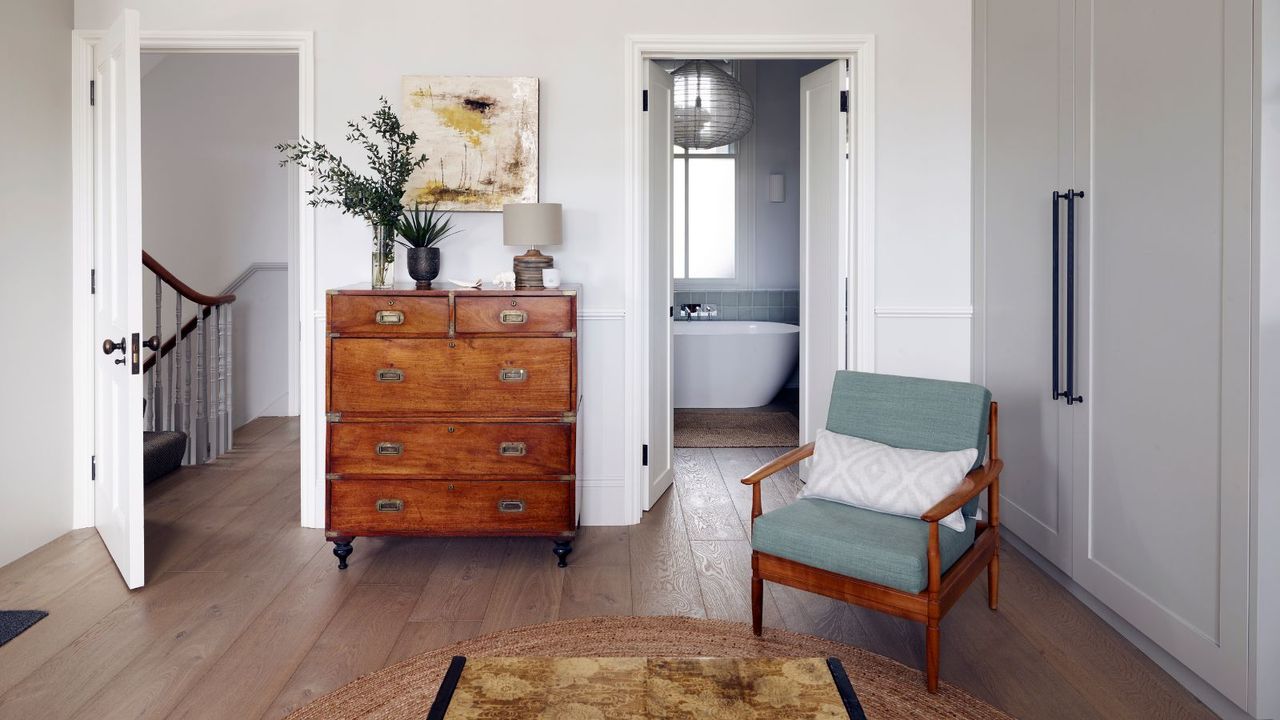 A white bedroom with a brown chest of drawers with plants on top, an artwork above, a blue and wooden chair and an open door to a bathroom behind