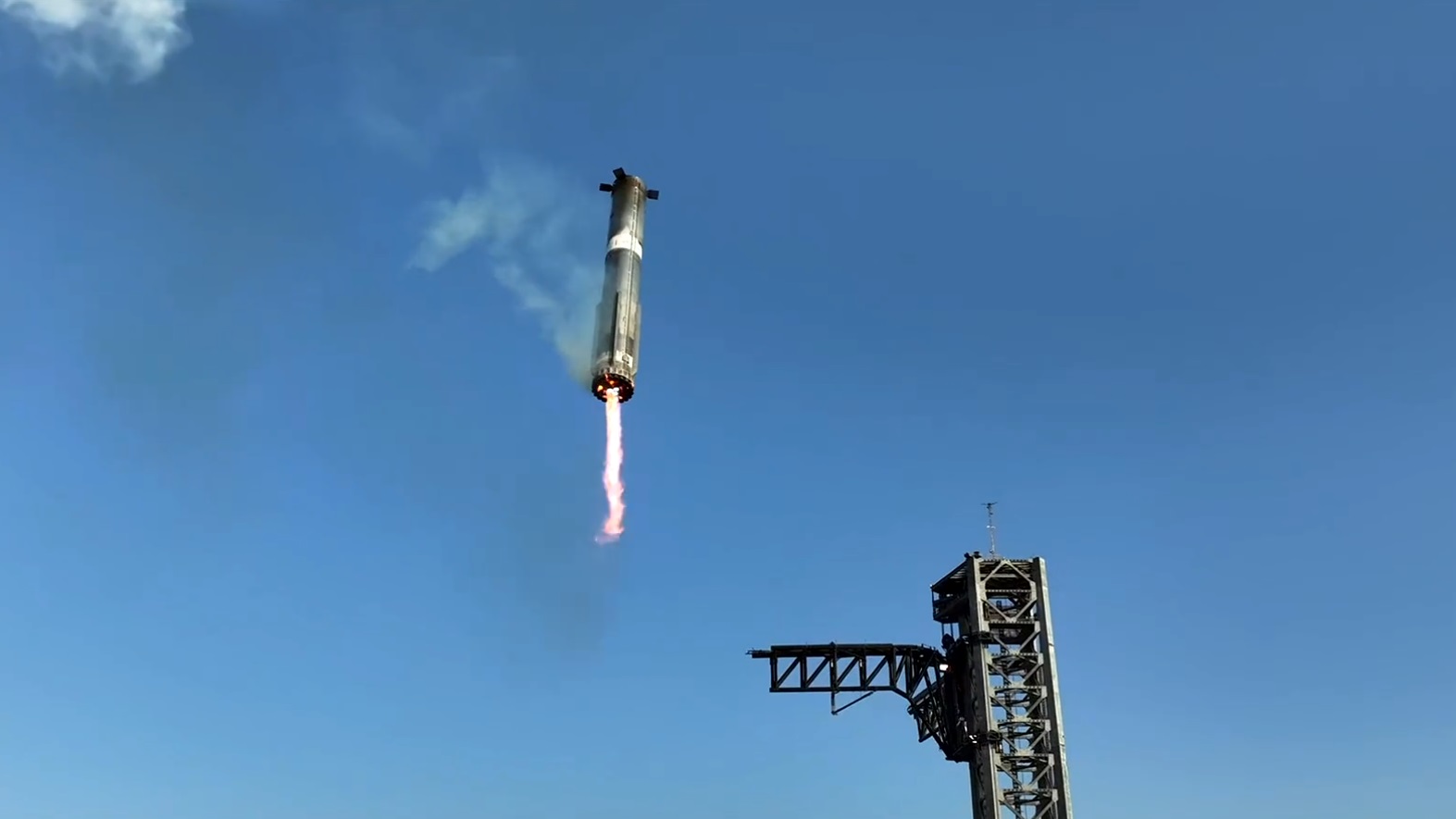 A giant silver SpaceX rocket booster hovers near its launch pad arms before landing