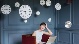 women sat in blue panelled room on dark red chair with laptop on lap and clocks floating around her head