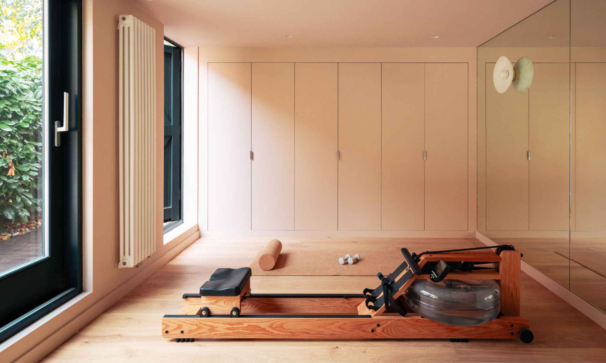 Wooden rowing machine on light hardwood floor, pink walls, built-in wardrobes, mirror, cork yoga mat, dumbbells.