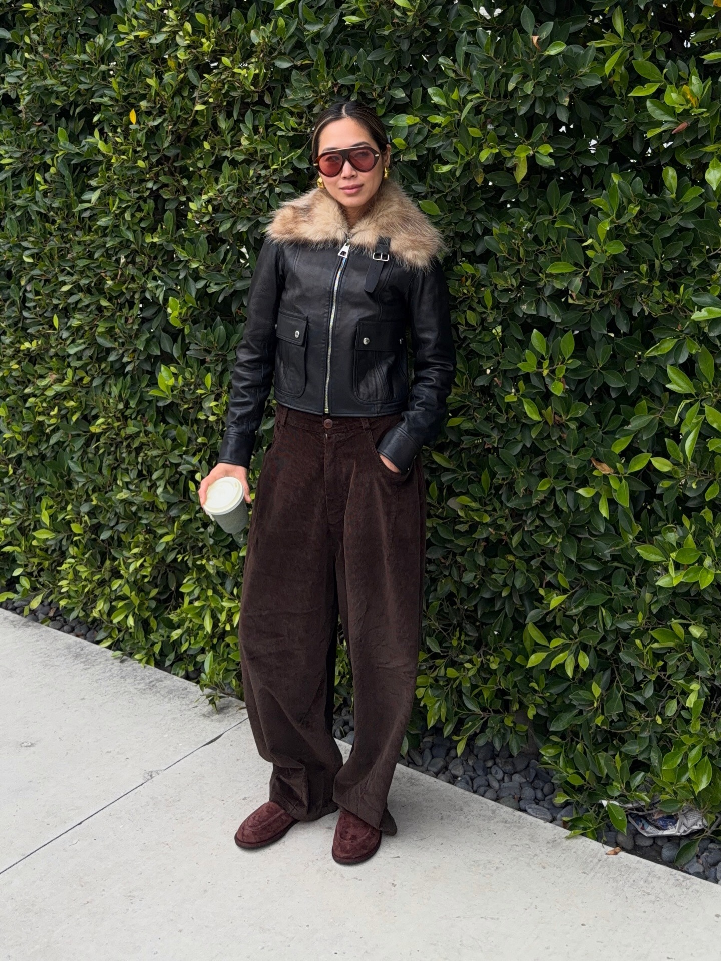 Aimee Song wears aviator sunglasses with red-tinted lenses, a leather biker jacket with a fur collar, brown corduroys and brown loafers.