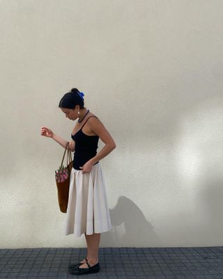 Sasha Mei wearing a black camisole with an off-white A-line skirt and Mary Jane flats.