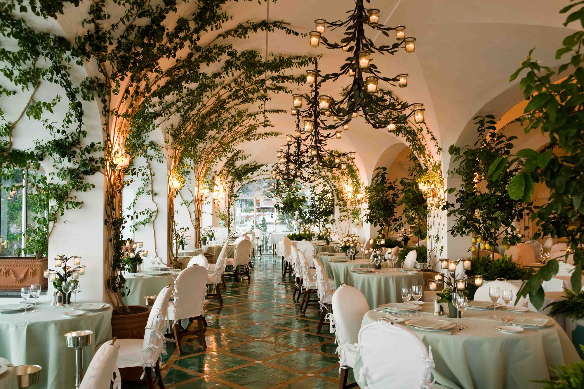 A romantic restaurant terrace with white-washed, arched walls, climbing plants decorated with candles, turquoise tiled floors with paler matching table cloths, and white fabric-covered chairs.