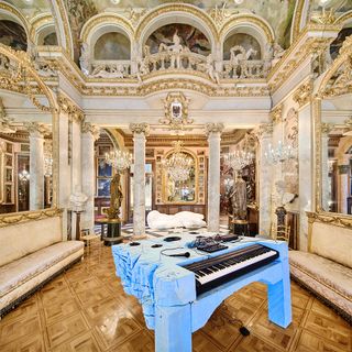 A grandly designed gold and cream room with Chandeliers and bold gold designed and pillars and a piano with blue chopped up foam placed in the center of the room