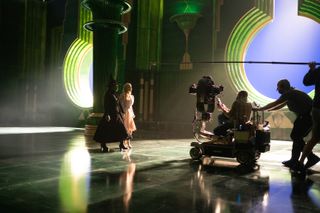 Two actors (Cynthia Erivo as Elphaba and Ariana Grande as Glinda) are filmed as they walk towards a camera and boom mic rig on an Emerald City set, in 'Wicked Part One.'