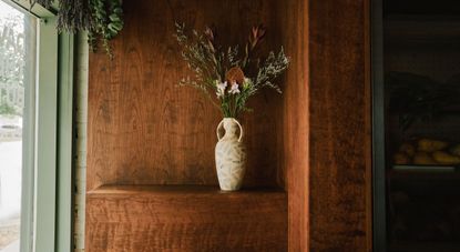 A wood-clad cafe, bakery, and butcher decorated with a built-in shelf topped by a ceramic vase with raw flowers is illuminated by natural light and boasts a pale green-painted facade.