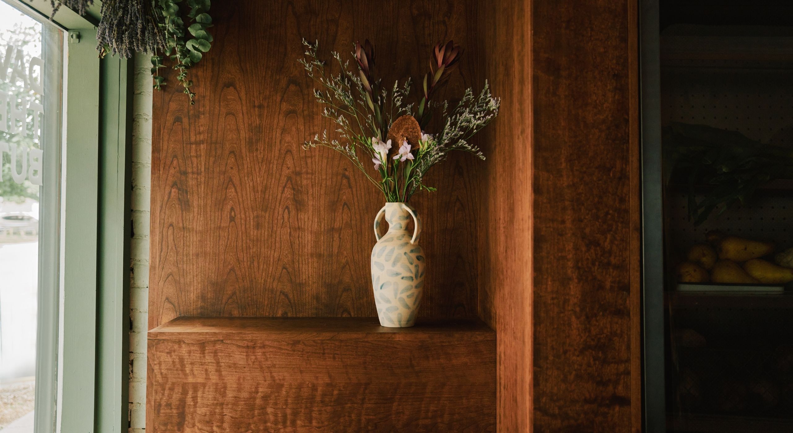 A wood-clad cafe, bakery, and butcher decorated with a built-in shelf topped by a ceramic vase with raw flowers is illuminated by natural light and boasts a pale green-painted facade.