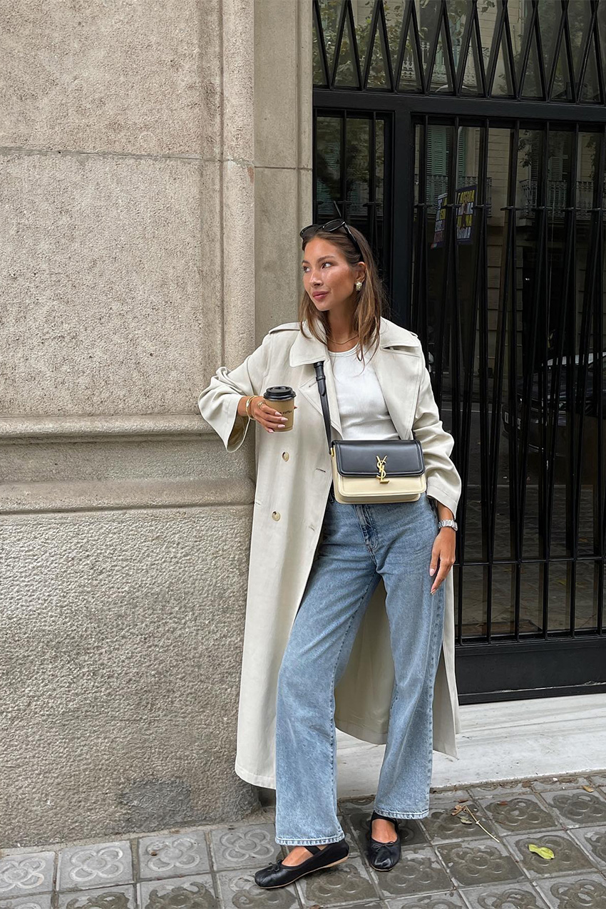 summer airport outfit is shown in a street style photo of a French woman standing outside wearing a tan trench coat over a white tank top with straight-leg jeans, black Miu Miu ballet flats, a two-tone Saint Laurent crossbody bag, black sunglasses, and gold earrings