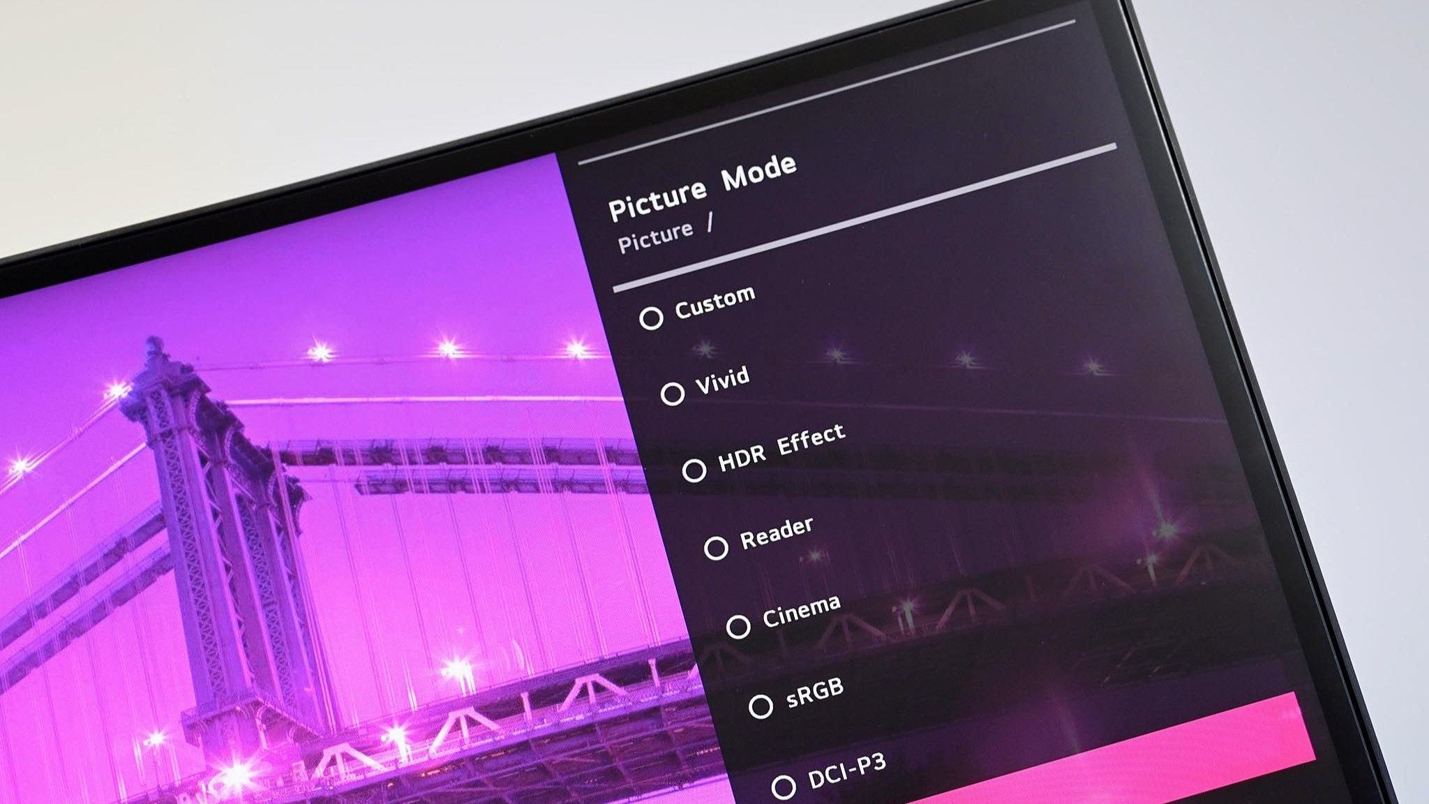 Angled close up of an LG PC monitor displaying an on screen menu for Picture Mode presets including Custom, Vivid, HDR Effect, Reader, Cinema, sRGB, and DCI-P3 over a purple cityscape background.