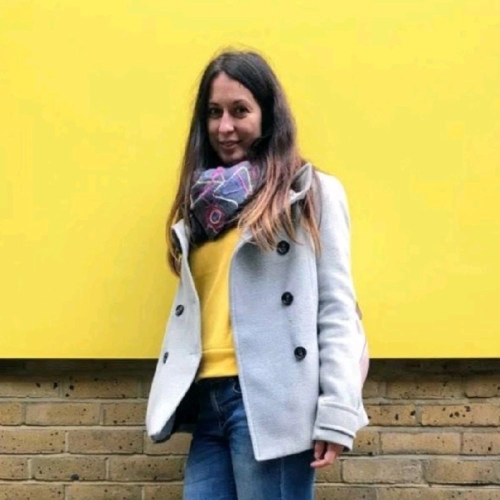 A brown haired woman in a grey peacoat standing against a yellow wall and smiling