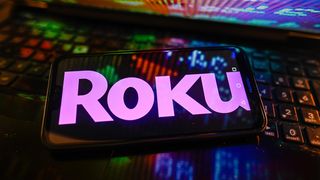 The Roku logo on a smartphone, resting on a laptop keyboard with an illuminated screen reflecting red, green, and blue light.