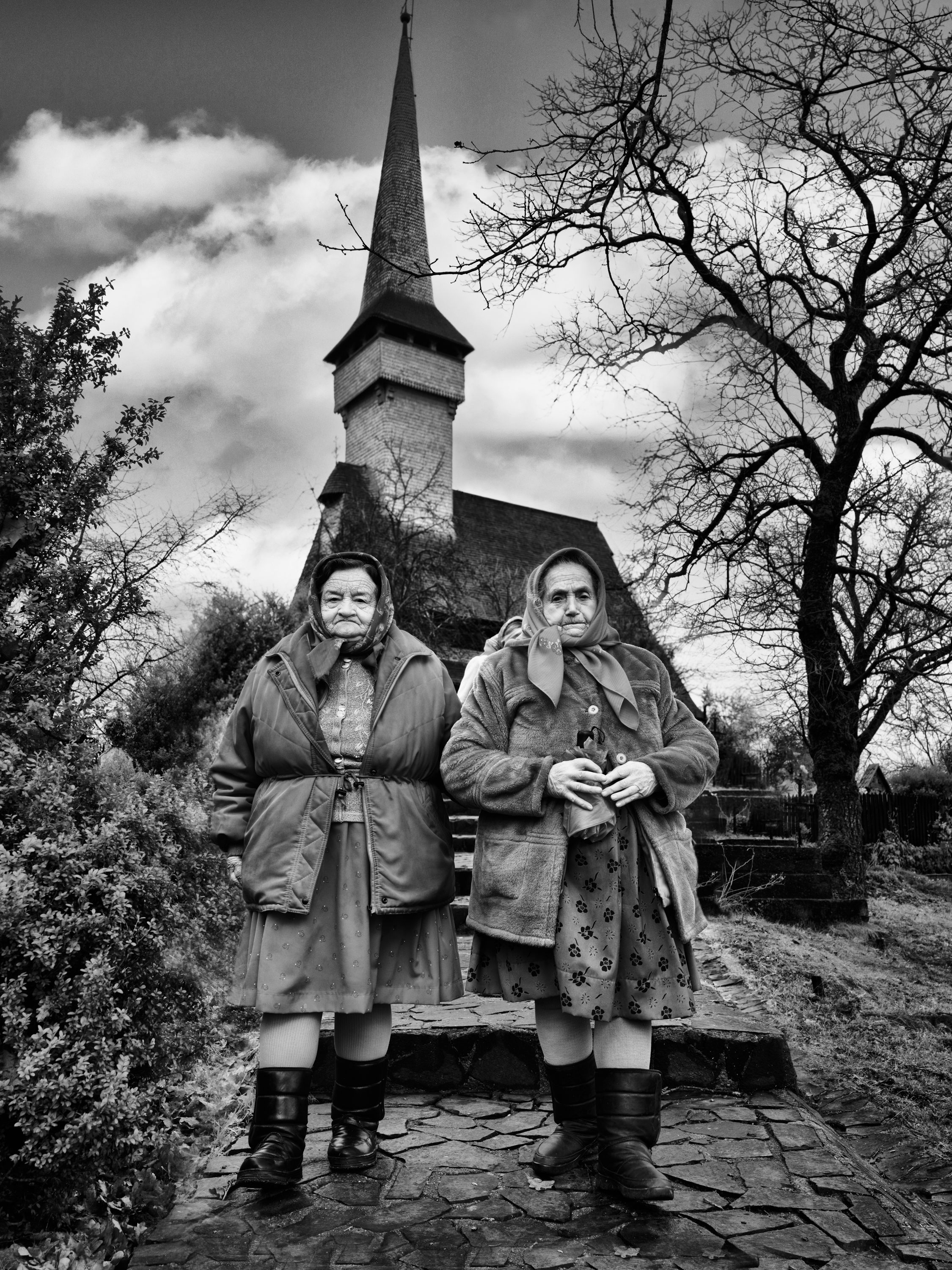 Black and white portrait of two elderly women in headscarves and heavy coats standing on stone steps before a tall wooden church spire, framed by bare tree branches.