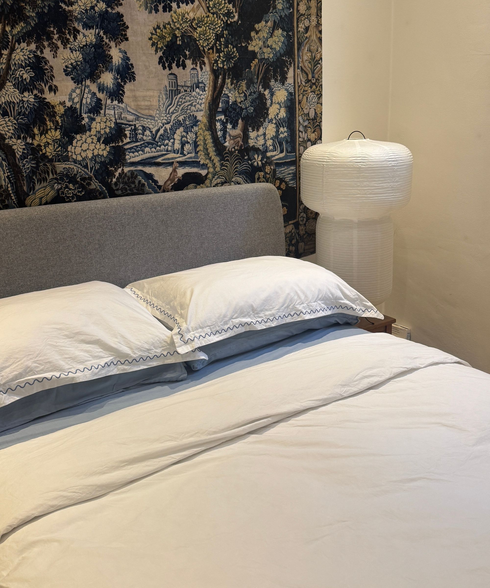 bed made with white christy bedding review with blue sheets, paper lantern, tapestry behind bed, and gray headboard