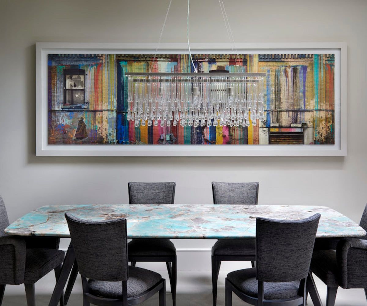 A kitchen-dining area with a painting on the wall and a small chandelier over a blue table and grey chairs