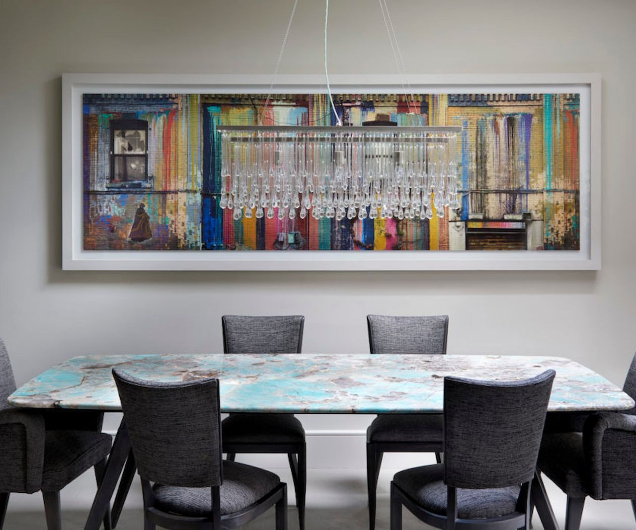 A kitchen-dining area with a painting on the wall and a small chandelier over a blue table and grey chairs