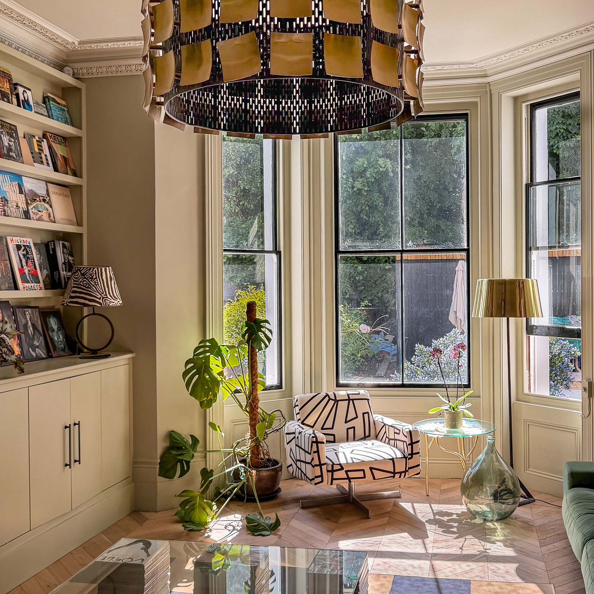 Bay window with floor lamp, armchair covered in abstract pattern, glass coffee table and houseplants by built in bookshelf area