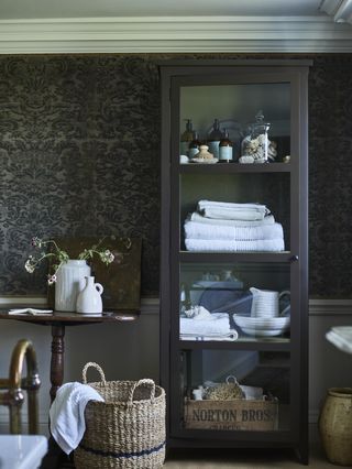bathroom with glazed freestanding cabinet , baskets, side table, bath in foreground, wallpaper