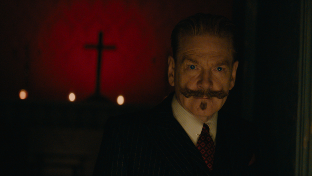 Kenneth Branagh stands eerily in front of a red background displaying a cross in A Haunting in Venice.