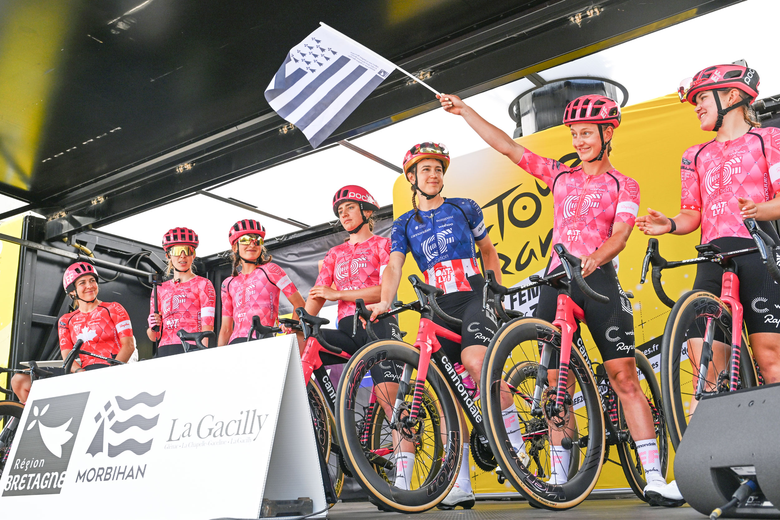 A low-angle shot of Breton rider Cédrine Kerbaol standing on stage and waving the black-and-white striped Breton flag alongside her EF Education-Oatly teammates before stage 3 of the 2025 Tour de France Femmes