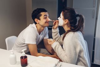 Home date ideas: Woman applying clay mask on her boyfriend's face. Young loving couple taking care of skin at home