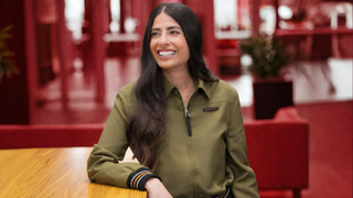 Asha Sharma sits at a wooden table, wearing a green jacket, while the blurred background of a modern office space is tinted in a deep red hue.