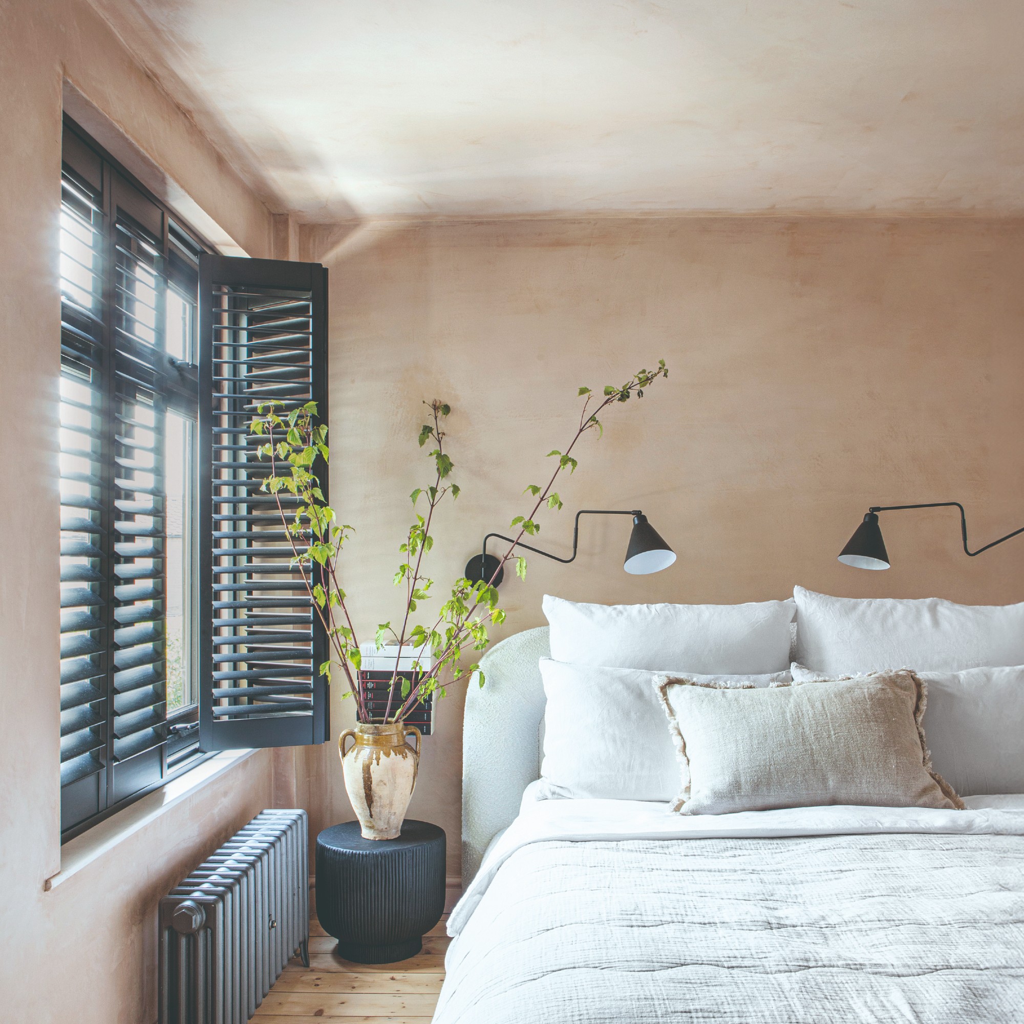 A limewash-painted bedroom with contrasting black window shutters and radiator