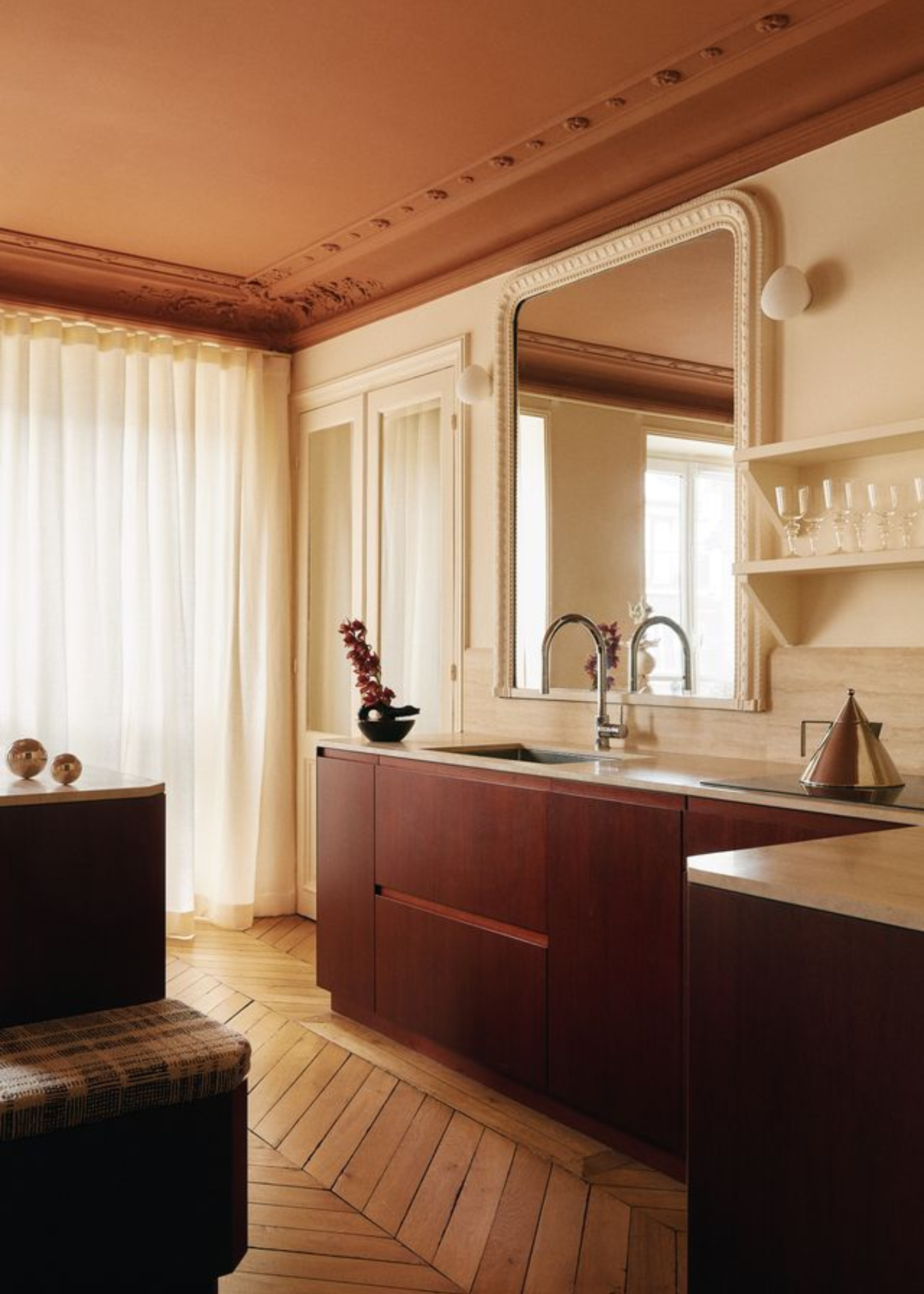 A beautiful open concept kitchen with moulding around the ceiling and the mirror, a triangular kettle, a shelf of glasses, round wall sconces, a vase with a flower stem, and wood flooring