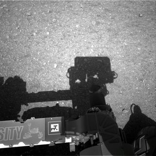 This is the first image taken by the Navigation cameras on NASA's Curiosity rover. It shows the shadow of the rover's now-upright mast in the center, and the arm's shadow at left.