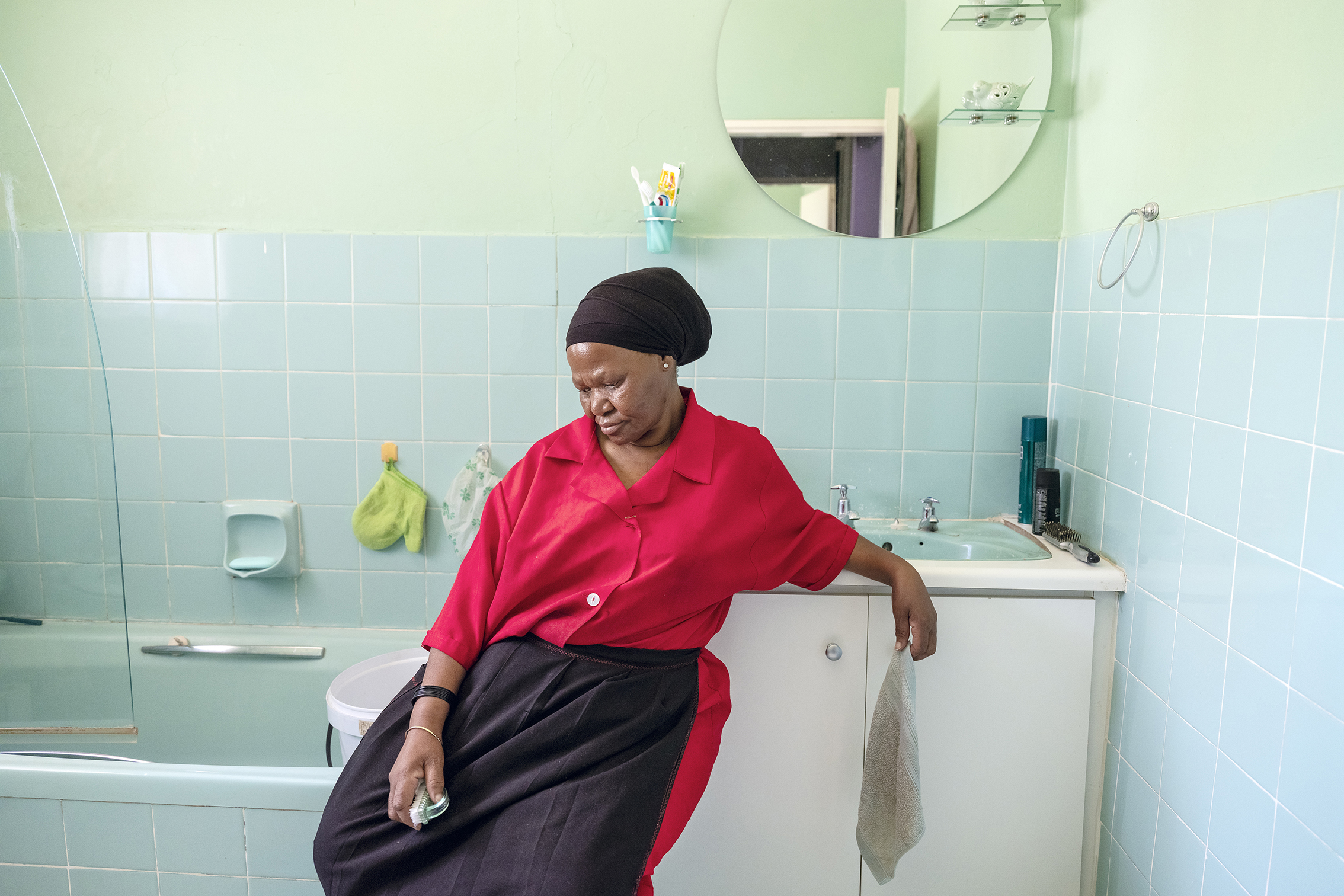 Color photograph of a woman sat in a bathroom looking down. She holds a sponge in her hand