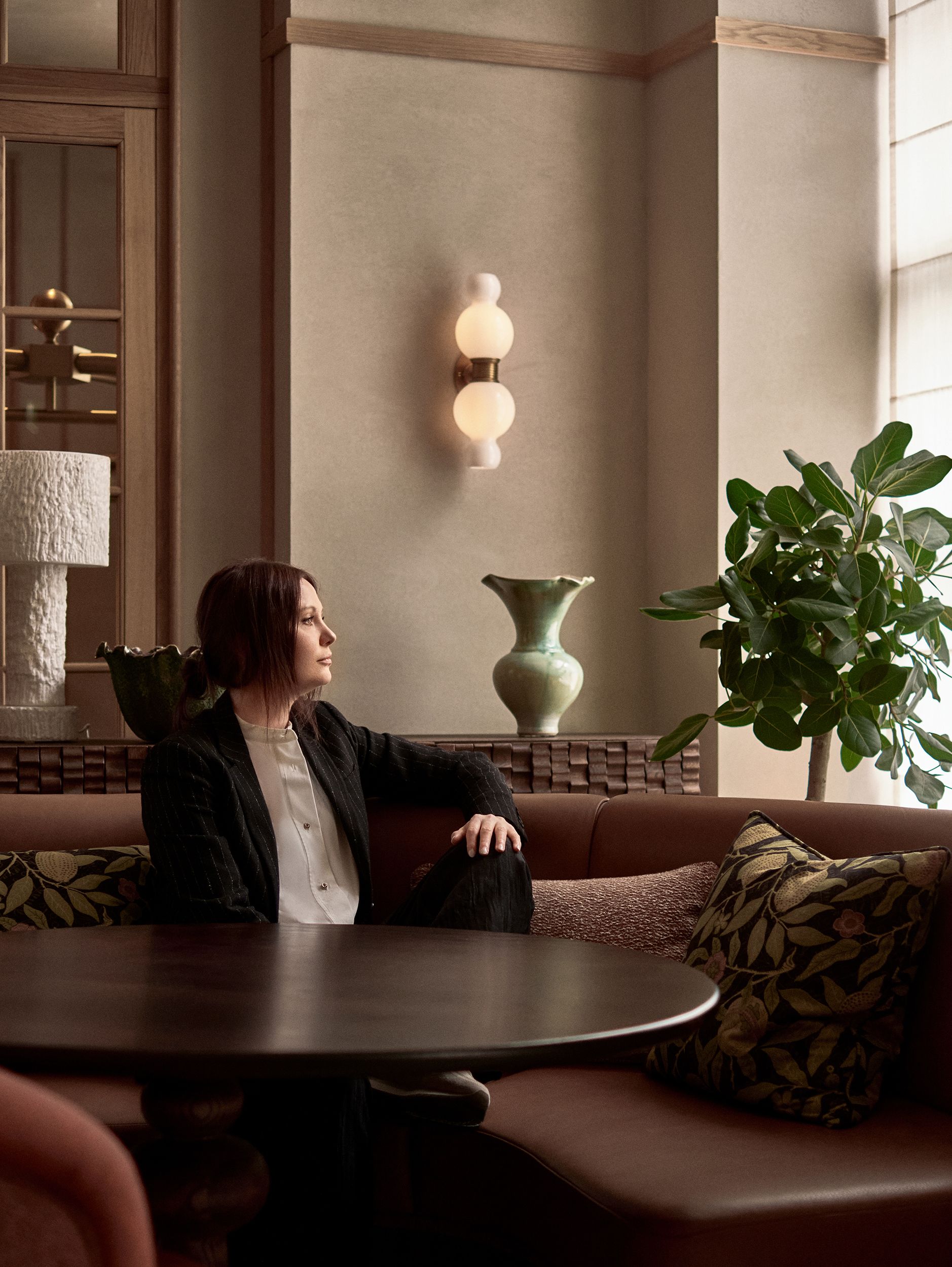 A young woman dressed in a black blazer and white Korean-style shirt stares out of a window while sitting on a chocolate brown leather banquette immersed in patterned fabrics, plants, sculptures, and soft light.