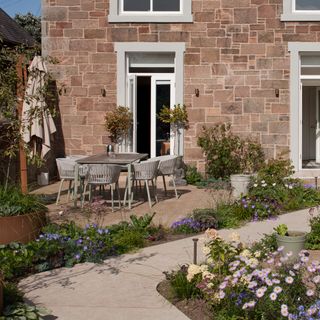 a dining table, chairs and parasol in front of a house with a winding pathway and plenty of purple flowers