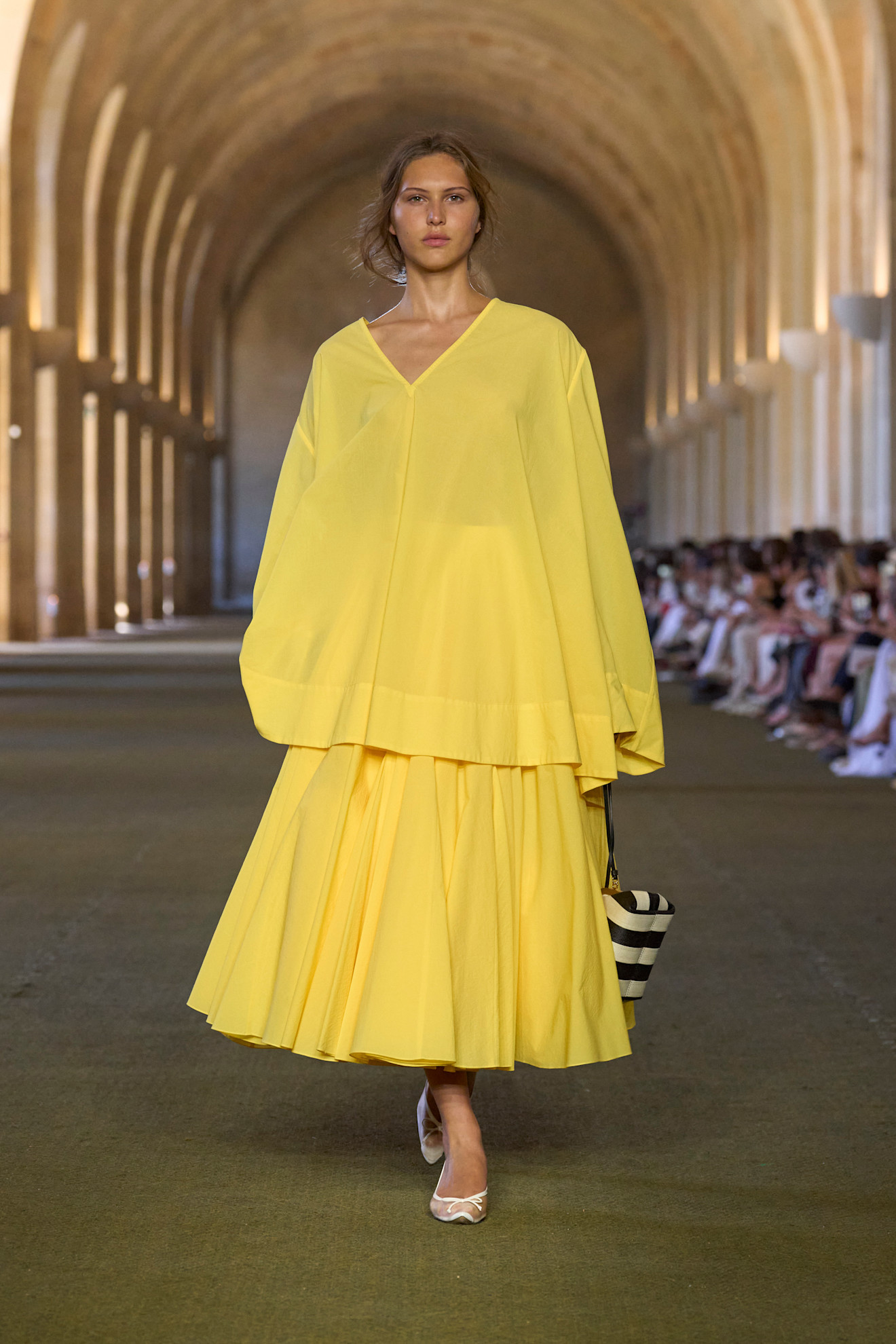 A Jacquemus model wearing the citrus yellow color trend in an oversize v-neck yellow blouse styled with a matching voluminous pleated midi skirt, white mesh flats, and a black and white striped clutch bag.