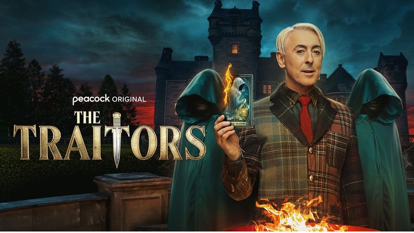Host Alan Cumming dressed in a tartan jacket holding a flaming portrait of a traitor as he stands against the gloomy backdrop of Ardross Castle, and flanked by two hooded, ominous figures in green cloaks. 