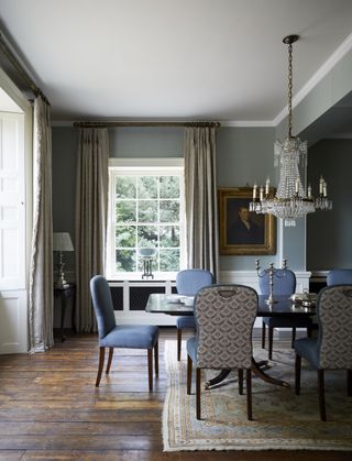 grand dining room with grey/blue walls, chandelier, large mahogany table, upholstered blue chairs, wooden floors, table lamps, artwork, rug