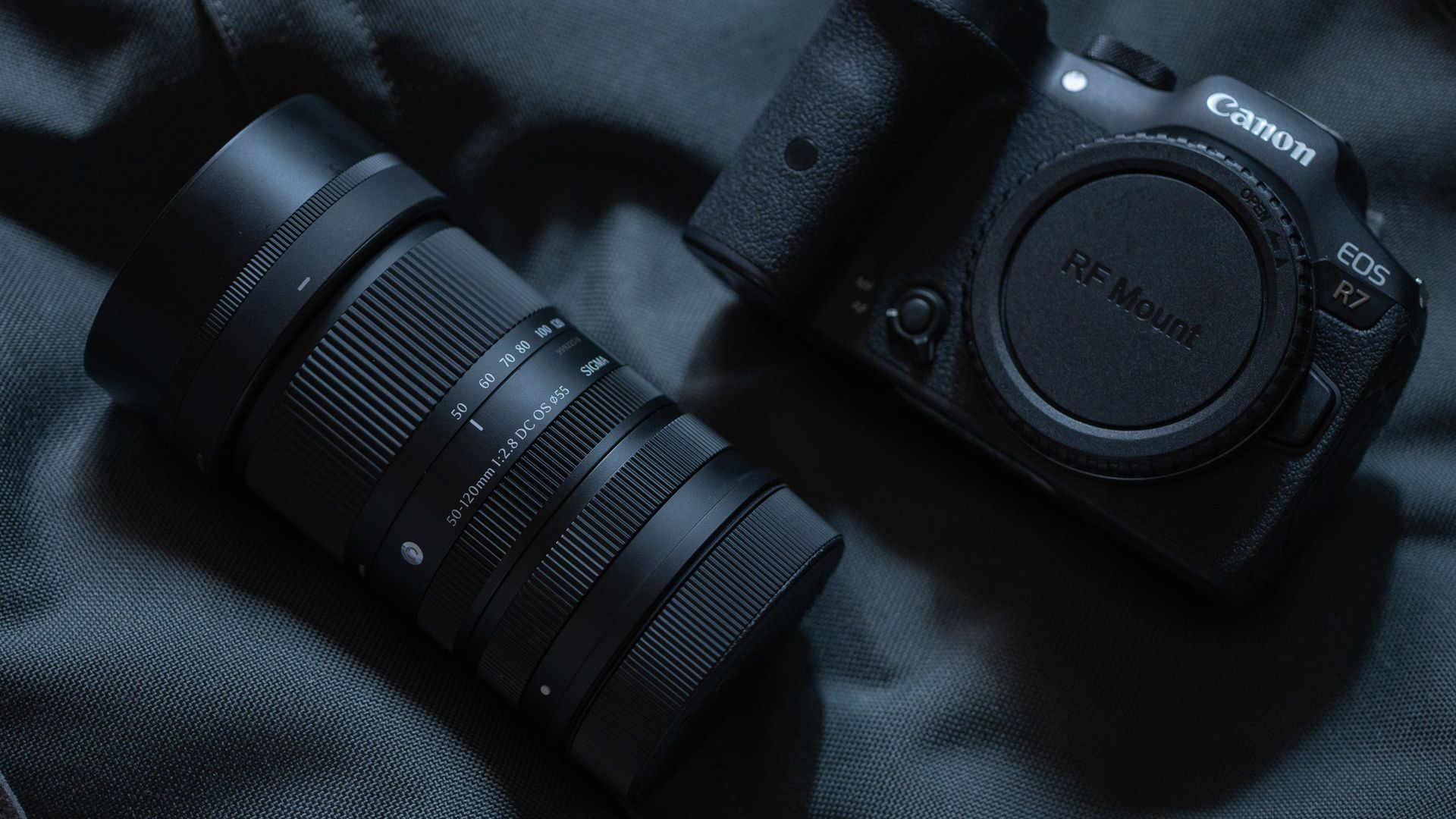 Sigma 50-120mm F2.8 DC OS Contemporary lens next to a Canon EOS R7, in a lowkey lit studio with dark background