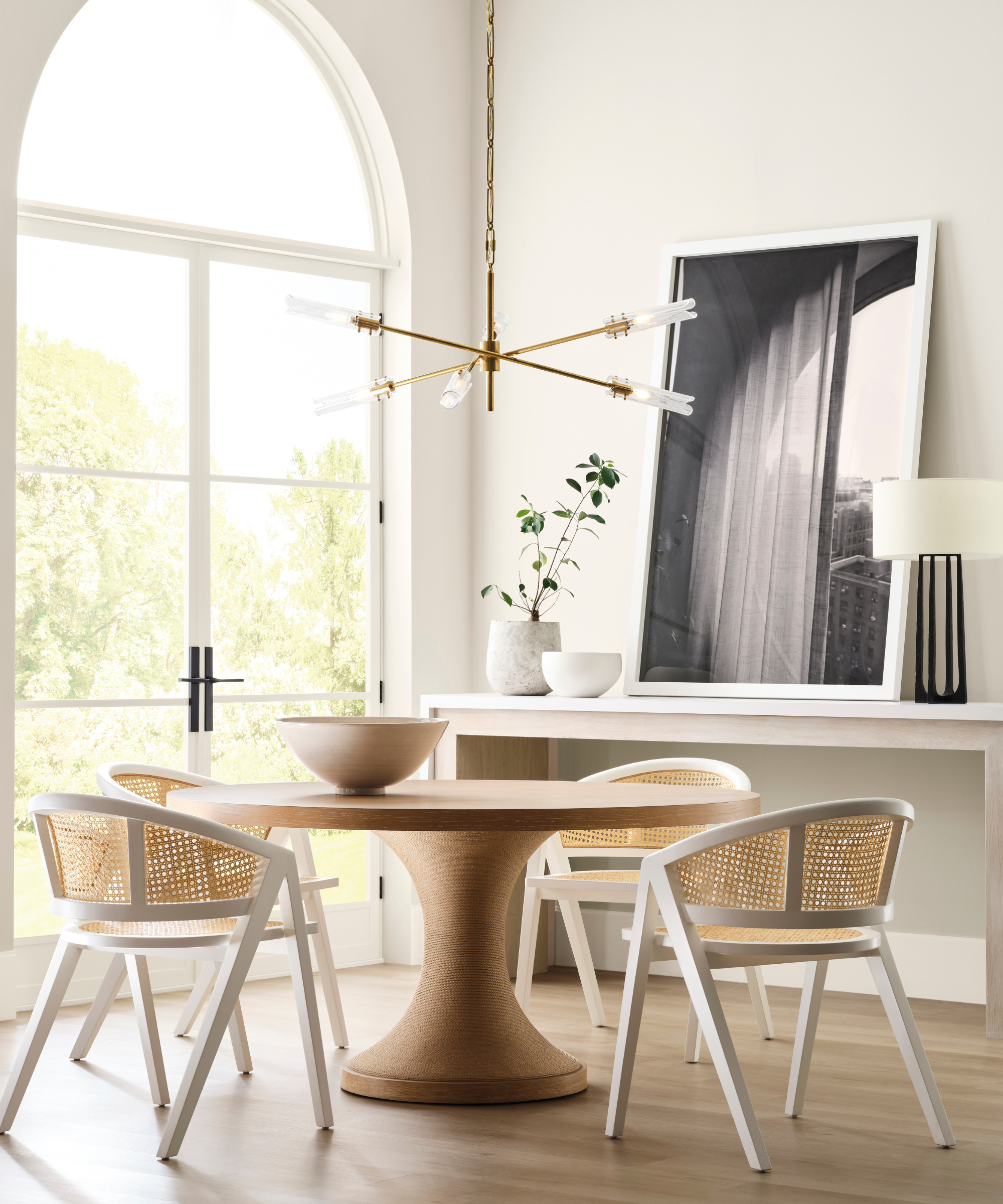 A bright dining room with high ceilings, a modern chandelier, a round wooden table, and four wicker chairs