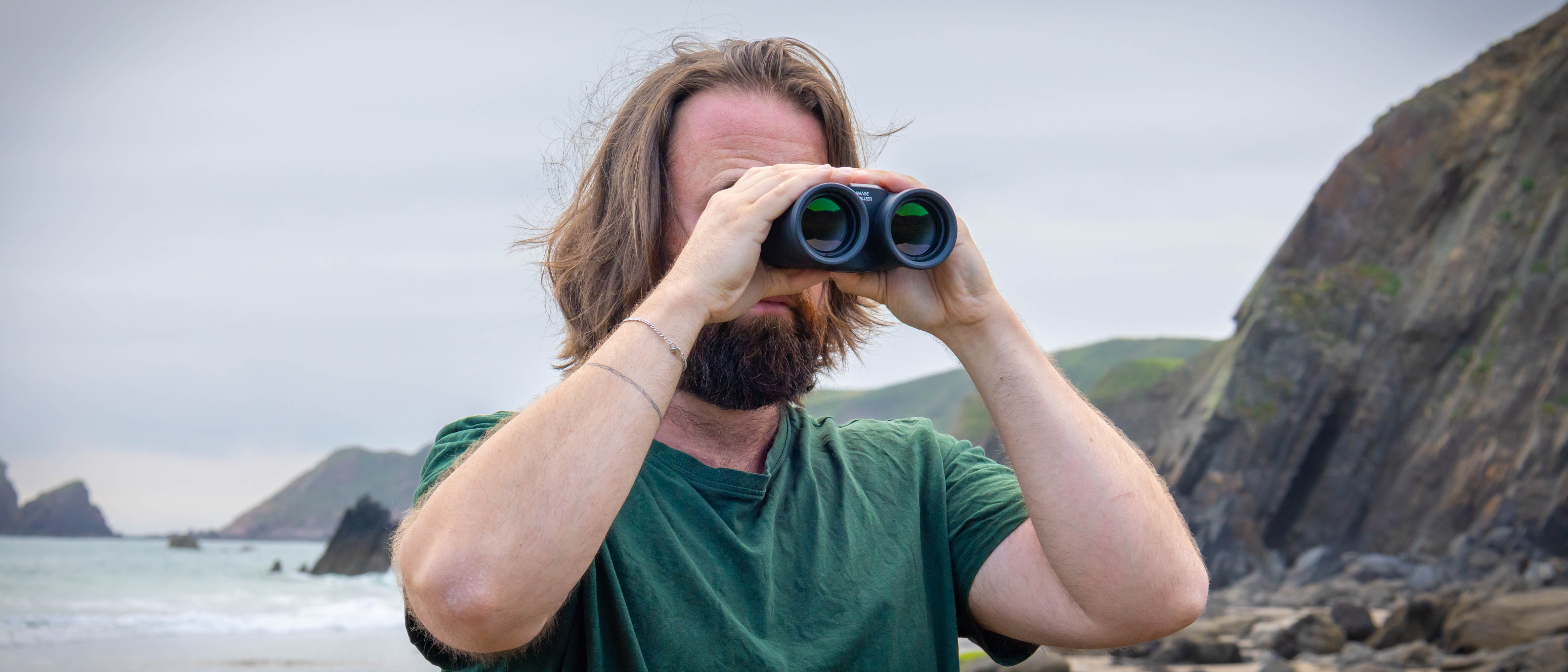 A male using the Canon 12x36 IS III binoculars with the coastline and sea behind them.