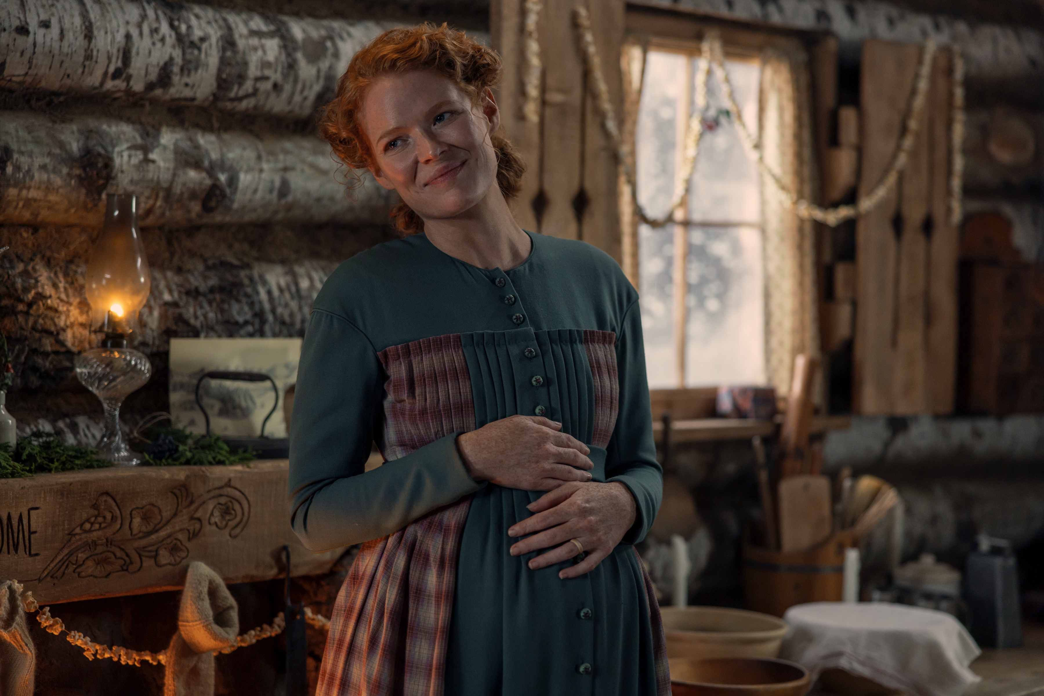 Actress Crosby Fitzgerald as Caroline Ingalls smiles with her hands resting on her belly, while standing in a 19th century frontier home, in Netflix's 'Little House on the Prairie.'