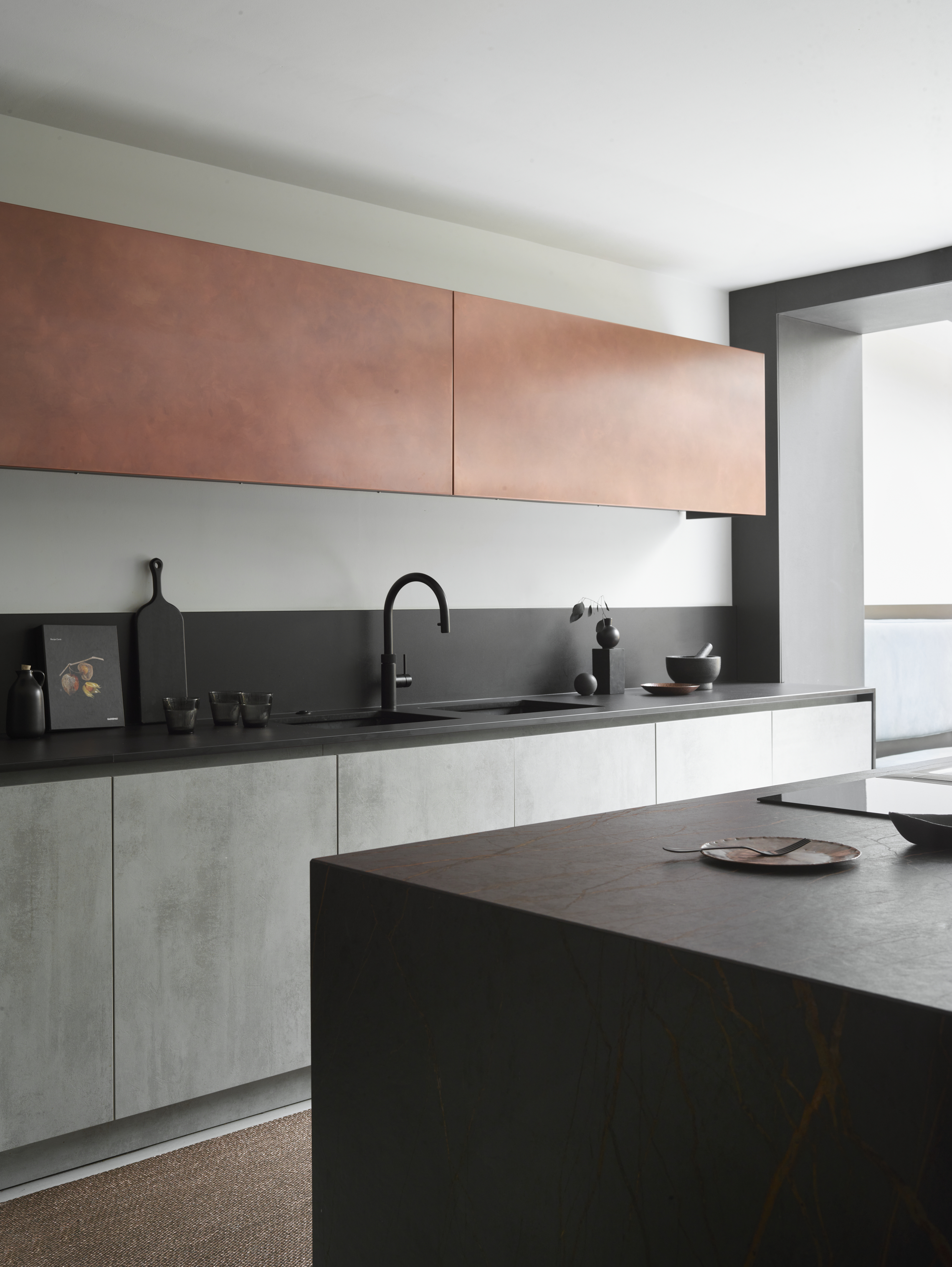 A kitchen with grey under cabients, a black counter and a brass upper cabinets, all without handles or knobs