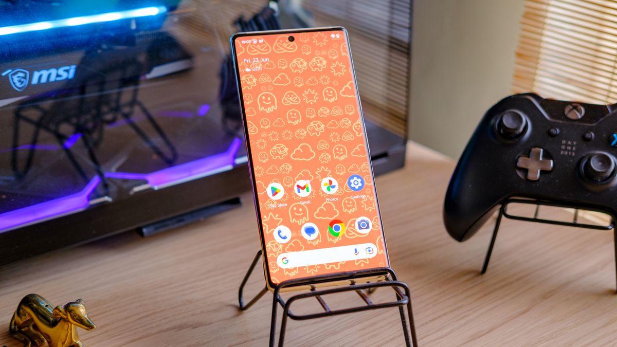 A photo of a Google Pixel 6 Pro with an orange emoji wallpaper, on a phone stand with a gaming computer in the background emanating a purple and blue glow