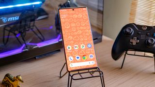 A photo of a Google Pixel 6 Pro with an orange emoji wallpaper, on a phone stand with a gaming computer in the background emanating a purple and blue glow